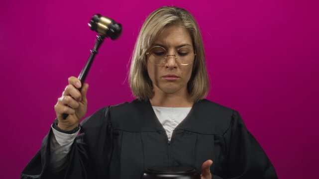 Blonde woman in judge robe holds gavel with raised arm in magenta studio; justice authority seriousness.