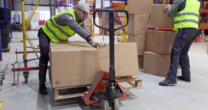 Workers loading boxes with pallet truck in warehouse. Two workers in safety vests move parcels and packages for delivery, shipping and storage. Concept of efficient warehouse logistics teamwork.