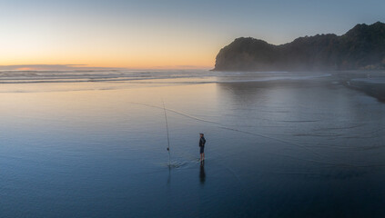 A lone fisherman stands in the surf with his fishing pole at sunset in Piha, Auckland, New Zealand. He is hoping to catch fish for dinner for his family. © Zenstratus