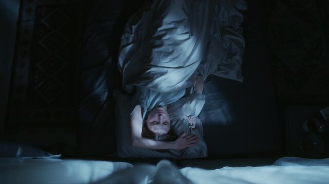Top view of young adult woman tossing and turning in bed trying to get to sleep then nervously throwing away pillow during night in bedroom