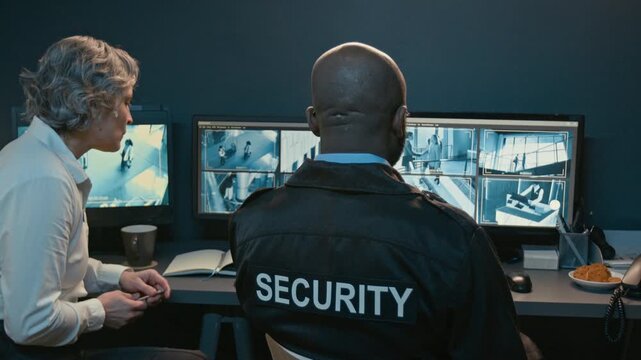 Back view shot of two people watching CCTV footage on multiple computer monitors while coordinating surveillance at night with focus on word Security printed on uniform with copy space