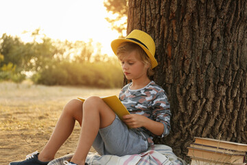 Fototapeta premium Cute little boy reading book near tree in park