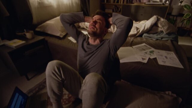 High angle shot of adult man sitting on bedroom floor holding head with hands while struggling with intense anxiety attack in dimly lit room, shaky camera