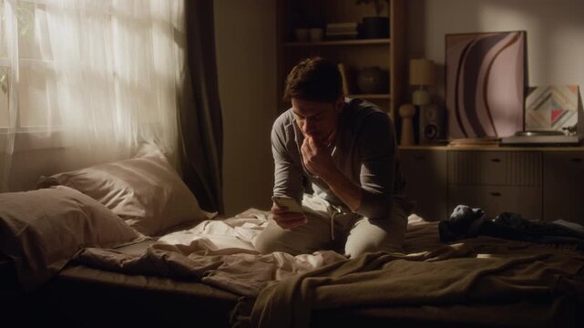 Full length shot of nervous adult man sitting on bed with legs tucked under, scrolling through bad news on phone in dimly lit bedroom then clutching head in despair