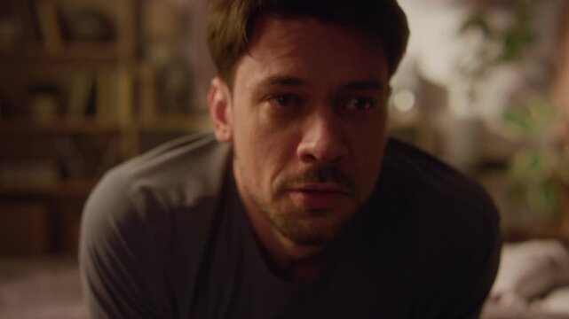 Close up on adult man clutching chest in fright struggling with panic thoughts swaying back and forth while trying to calm down with glass of water in dimly lit bedroom