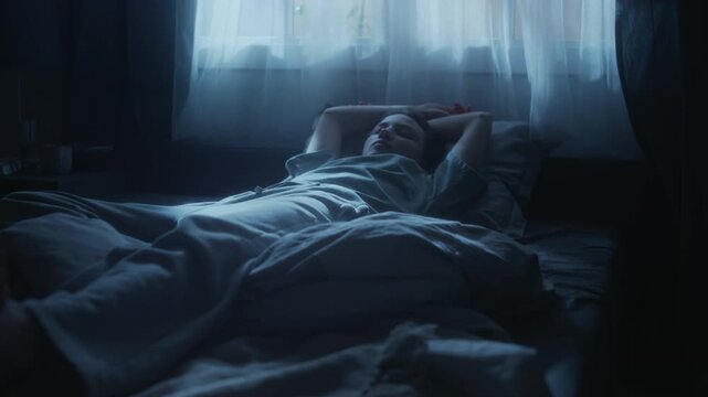 Rack focus of young adult woman lying in bed in moonlight trying to fall asleep, stretching arms then turning to other side in dark bedroom