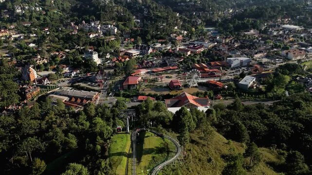 Aerial view of cable car gondolas and chair lift with people riding over forest hillsides at Capivari amusement park with roller coaster tracks and ferris wheel in Campos do Jordao, Brazil. Tilt down 