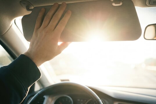 Driver raising a hand to pull down the grey sun visor in a car interior against bright glaring sunlight