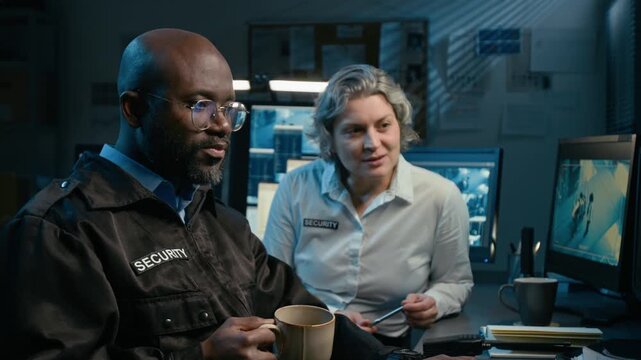 Medium shot of two security officers chatting and drinking coffee while watching CCTV feed on multiple computer monitors during night shift in office, scene lit by blue light with copy space