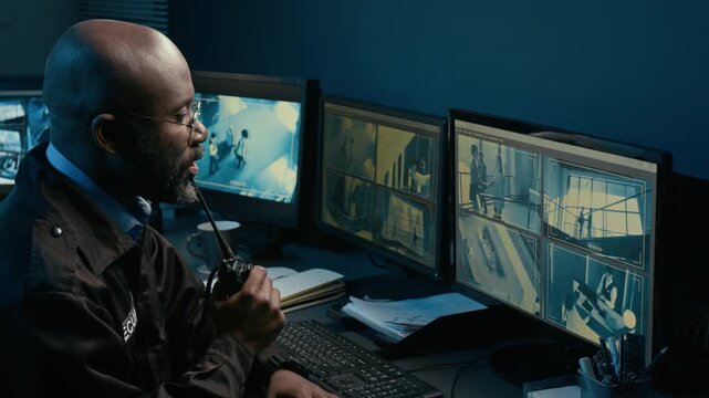 Rotating shot of Black man as security officer and giving commands speaking into portable radio while watching CCTV footage on multiple computer monitors in control room during night shift, copy space