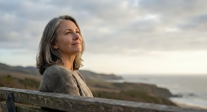 Mature blonde woman in cozy sweater enjoying peaceful moment on coastal bench at sunset, expressing serenity and contentment with ocean view backdrop.