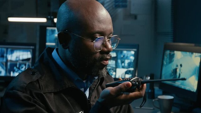 Tilt up shot of African American man speaking into portable radio while watching CCTV footage on computer monitors during night shift in security and surveillance office, copy space
