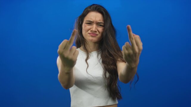 Teenage woman flips both middle fingers in studio while wearing a white top with furrowed brow; defiance anger rebellion frustration.