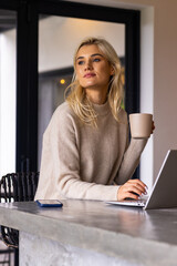Woman 20s typing on laptop at concrete counter holding mug in beige sweater blue-cased smartphone