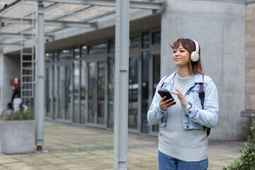 Woman standing at modern entrance wearing headphones with backpack using smartphone, copy space © wavebreak3