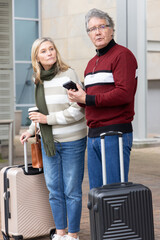Naklejka premium Senior couple waiting at station wearing red sweater green scarf holding suitcases phone coffee cup