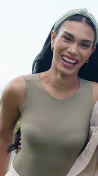 Vertical video: Laughing woman arm around friend reacting in park wearing olive top and headband