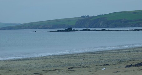 Naklejka premium Showing sandy beach pebbles and seaweed, shallow waves lapping at coast, rocks, grassy cliffs