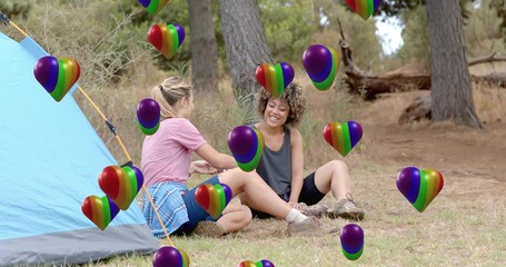 Sitting two friends chatting, smiling at campsite, with tent, plaid shirt, rainbow heart balloons