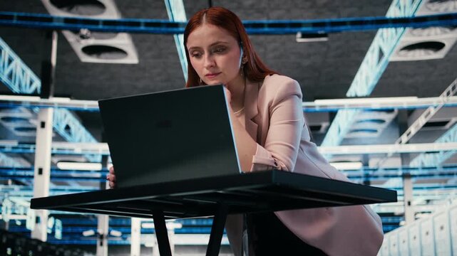 Female programmer at server room workplace desk using laptop to enhance operational effectiveness of equipment. System administrator in data center using notebook to monitor rackmounts, camera A