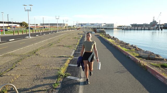 Young athletic woman in sportswear walking along a waterfront path in Alameda, California, holding a water bottle and a jacket after her morning workout with the USS Hornet in the background