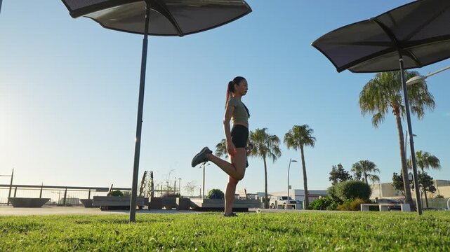 Fit young woman doing stretching exercises on a green lawn in a city park on a sunny day with palm trees in the background, preparing for her morning workout and focusing on her well-being