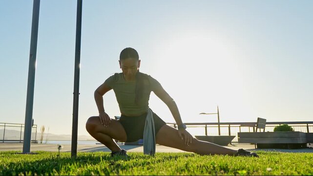 Fit young woman performing a side lunge stretch on a grassy park area by the shore, warming up her muscles before a run as the sun shines brightly in the background