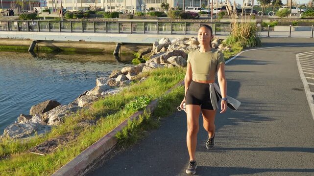 Fit woman walking along a waterfront path in a city park after her morning exercise, holding a water bottle and a sweatshirt while enjoying the sunny weather and a healthy lifestyle