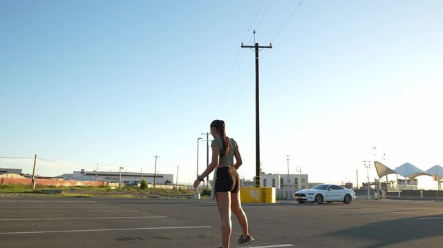 Young athletic woman with a ponytail preparing for her morning workout, stretching her legs in a large, empty parking lot near the shore with the bright sun shining behind her
