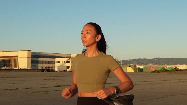 Fit asian woman in sportswear running on an old airfield during a beautiful sunset in Alameda, California, her determined expression showcasing her dedication to an active and healthy lifestyle