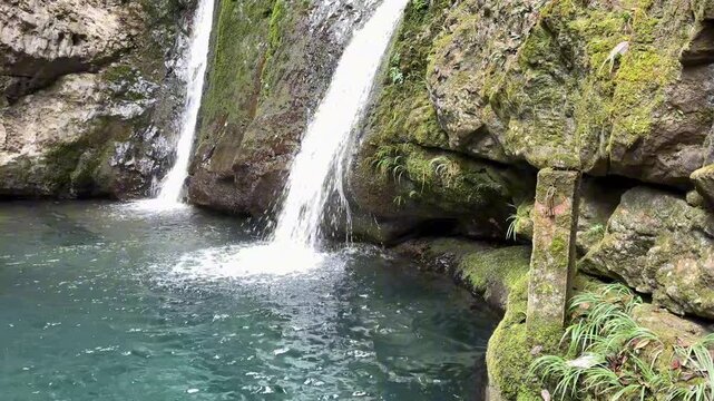 Elegant 4K 120fps shot of twin waterfalls flowing into a clear emerald pool, featuring mossy cliffs and a traditional stone pillar.