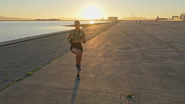 Athletic woman running on the spot and doing warm up exercises on a concrete path by the water, with the San Francisco skyline visible in the background during a beautiful golden hour sunset