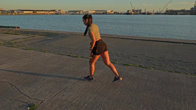 Young athletic woman performing warm-up exercises and stretching her legs on a concrete pier by the water at sunset, preparing for her evening run in an urban environment with a skyline view