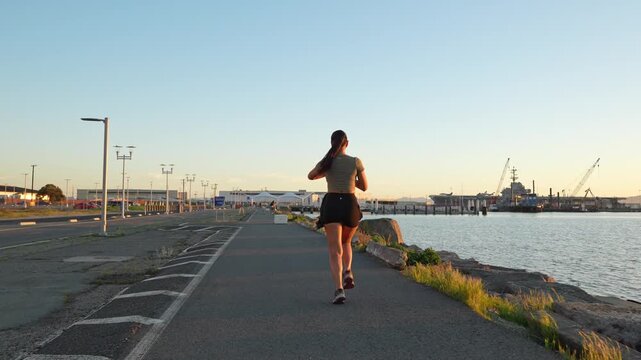 Young Asian woman jogging on a trail next to the ocean at sunset for her daily exercise, promoting an active and healthy lifestyle while enjoying the waterfront view of Alameda, California