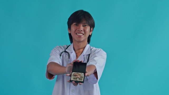 Young Asian male doctor in white lab coat smiling while presenting pair of hearing aids in case against blue background