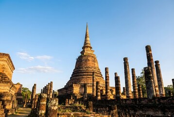 Obraz premium Majestic ancient Thai pagoda (Chedi) at Sukhothai Historical Park, ruins of stone pillars, serene atmosphere during golden hour, warm sunlight hitting the weathered brick texture, clear blue sky. 