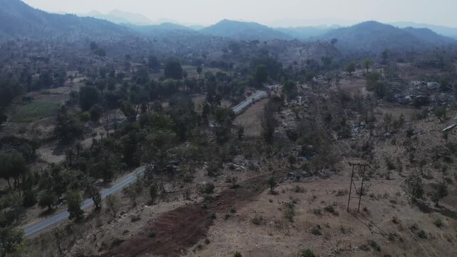 A white vehicle travels along a paved road winding through semi-arid hilly terrain with scattered trees and distant mountain ranges under soft atmospheric light in rural Rajasthan.