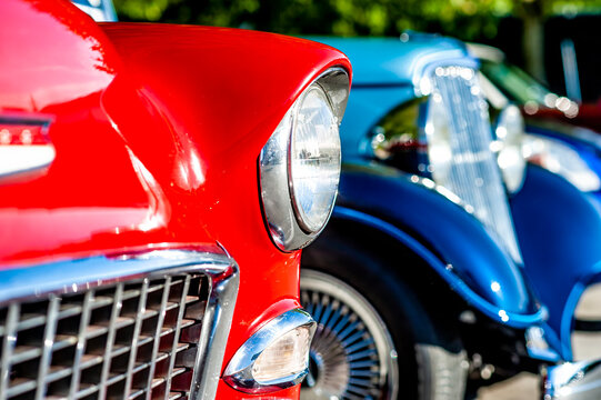Close up of a shiny red classic car with chrome grille and round headlight parked next to a blue vintage vehicle on a sunny day at an outdoor car show.