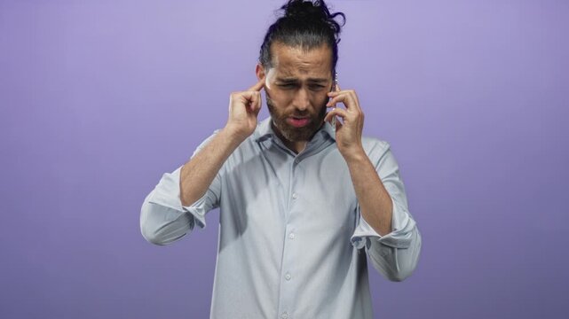 Young man with beard holding smartphone to ear while plugging ears with fingers in purple studio; frustration.