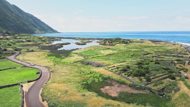 Drone ascent over Faja dos Cubres on Sao Jorge Island, Azores, Portugal. Coastal lagoon, terraced field and Atlantic shore road under bright sky