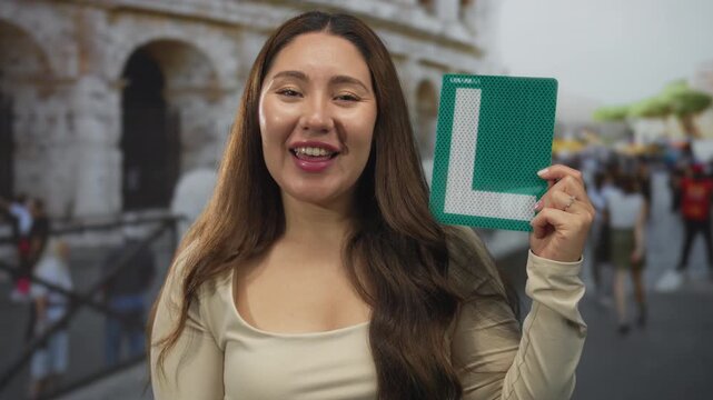 Woman in beige top holding green l plate pumps fist at coliseum building entrance under bright daylight; achievement.