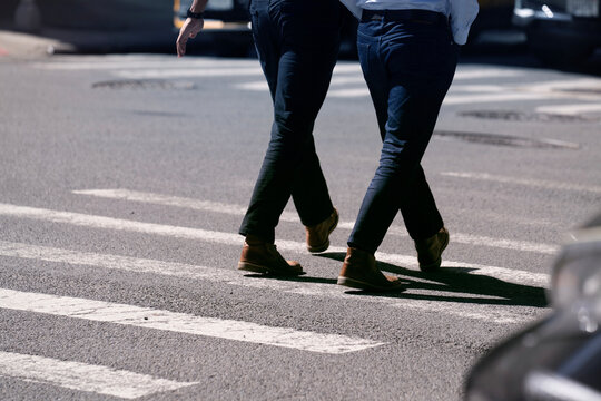 A low-angle candid shot captures two people walking across a street crosswalk. The focus is on their legs and feet, emphasizing movement and daily routine against the urban asphalt and white lines. It