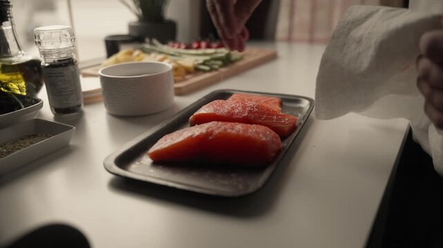 Two raw salmon fillets are placed on a tray while seasoning is sprinkled over them in a bright kitchen. Ingredients and vegetables are visible in the background during preparation of a home cooked mea