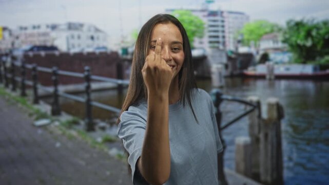 Woman showing middle finger directly to camera on a canal street in amsterdam wearing a light blue tshirt by a railing and boat; raw defiance.