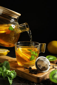 Pouring of tasty fruit tea from glass teapot into cup on black grunge table, closeup