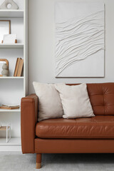 Fototapeta premium Brown leather sofa with soft pillows and shelving unit near white wall in living room