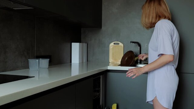 Woman in pajamas is washing dishes in the kitchen sink. After finishing the dishes, she bends down and starts cleaning the kitchen floor