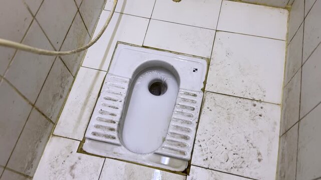 Top view of a ceramic squat toilet with flowing water on a stained tiled restroom floor, showing worn sanitation fixtures and poor hygiene conditions in an indoor washroom.