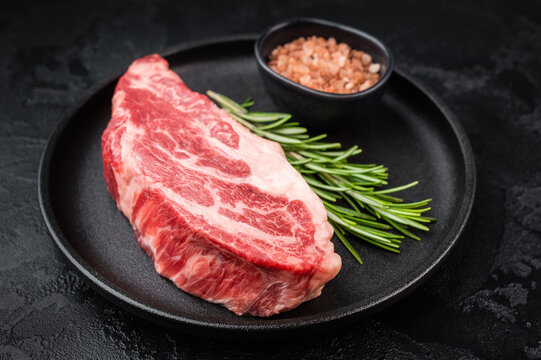Fresh raw beef chuck eye steak on black pan with rosemary and pink salt. Premium marbled beef meat for cooking and grilling.