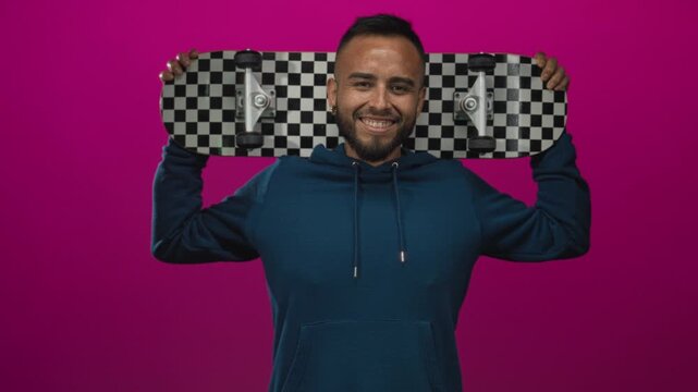 Young man smiling and holding checkerboard skateboard behind his head while wearing blue hoodie in studio magenta; playful joy.